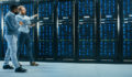 IT Technician with a Laptop Computer and Black Male Engineer Colleague are Talking in Data Center while Walking Next to Server Racks. Running Diagnostics or Doing Maintenance Work.
