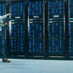 IT Technician with a Laptop Computer and Black Male Engineer Colleague are Talking in Data Center while Walking Next to Server Racks. Running Diagnostics or Doing Maintenance Work.