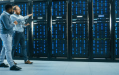 Principal Asset Management on data center expansion and evolution IT Technician with a Laptop Computer and Black Male Engineer Colleague are Talking in Data Center while Walking Next to Server Racks. Running Diagnostics or Doing Maintenance Work.