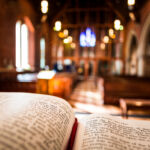 Bible open at the book of John inside an Anglican Church of England church. The focus is on the foreground of the bible, while in the background, defocused, are the pews and arches of the historic eighteenth century building. Horizontal colour image with copy space.
