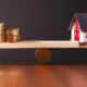 Image of a house and coins balancing on opposite ends of a wooden scale.